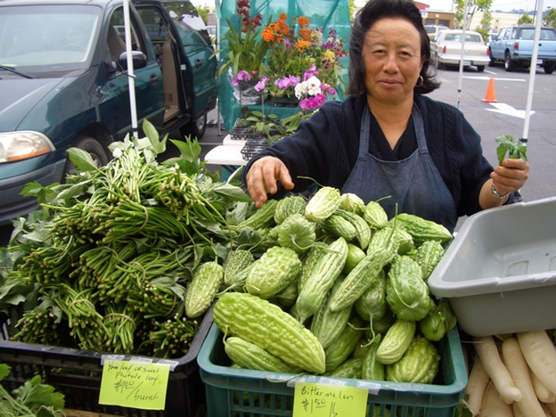 Hmong Farmer sells green produce in bins outdoors.
