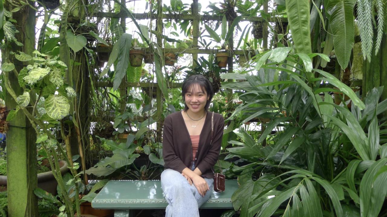 Photo of Emily Chung seated with one leg crossed smiling at the camera with a background of plants