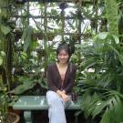 Photo of Emily Chung seated with one leg crossed smiling at the camera with a background of plants 